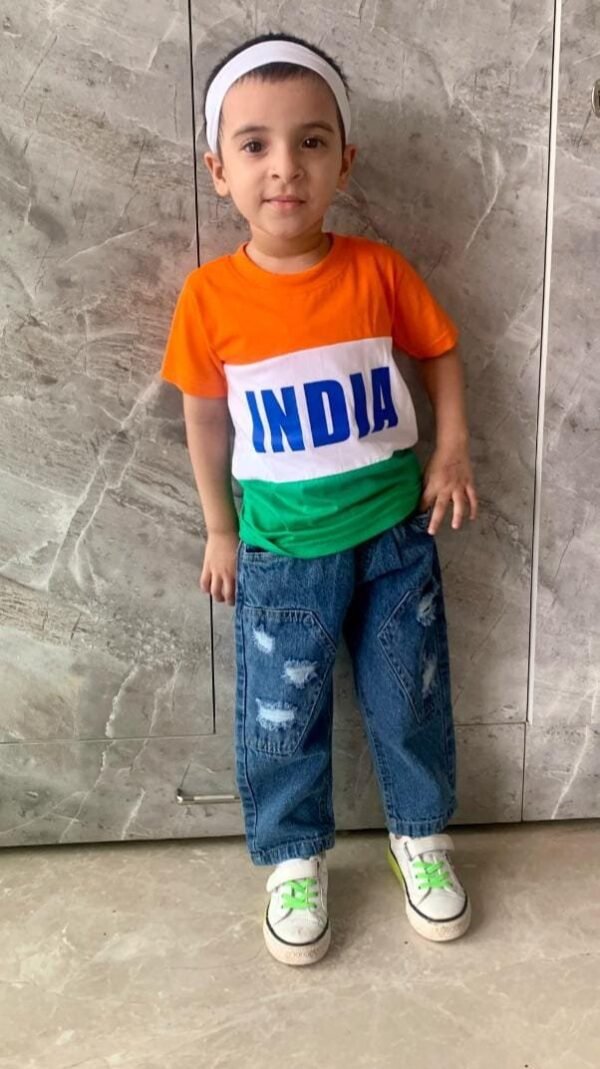 Child wearing a tricolor t-shirt with orange, white, and green stripes and “INDIA” written in blue, paired with distressed blue jeans and white sneakers with green laces, standing against a textured wall